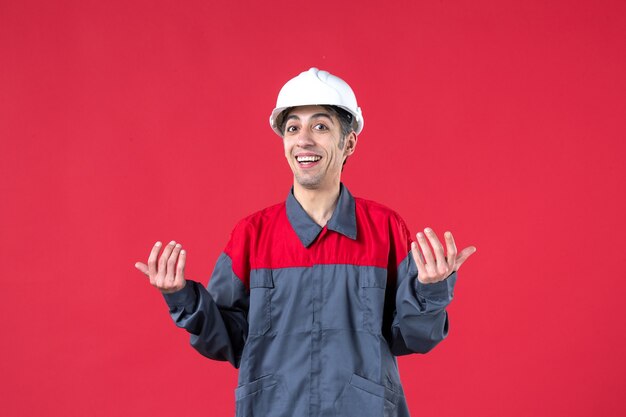 Colpo di mezzo corpo di giovane costruttore sorridente in uniforme con elmetto sul muro rosso isolato