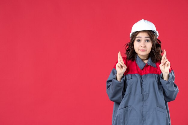 Colpo a metà corpo di un costruttore femminile incerto in uniforme con elmetto e incrociando le dita su sfondo rosso isolato