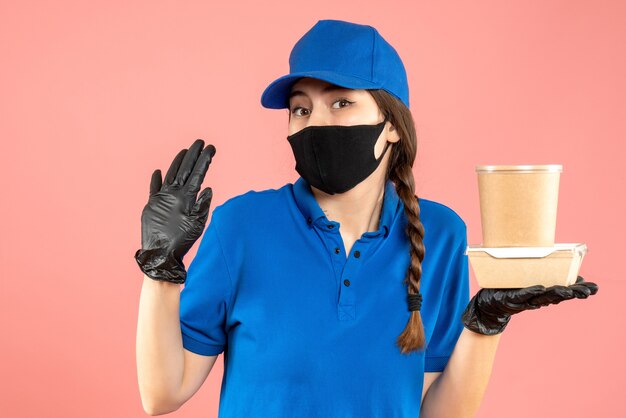 Half body shot of worried courier girl wearing medical mask and gloves holding small box coffee on pastel peach background