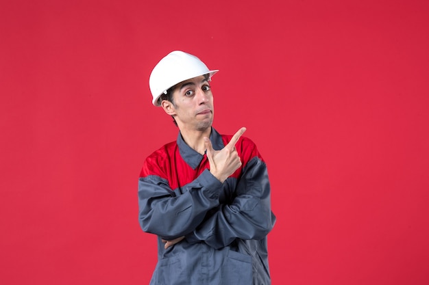 Half body shot of young builder in uniform wearing hard hat pointing up on isolated red wall