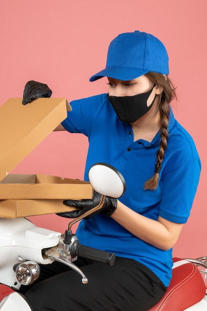 Half body shot of young curious female courier wearing medical mask and gloves sitting on scooter opening boxes on pastel peach background