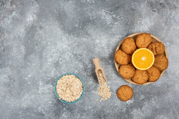 나무 판자에 홈메이드 쿠키를 넣은 반 자른 오렌지와 회색 테이블 위에 그릇에 오트밀.