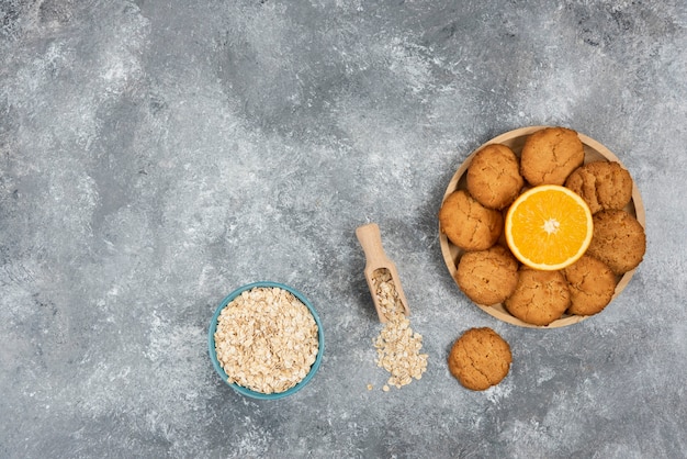 Half-cut orange with homemade cookies on wooden board and oatmeal in bowl over grey table.