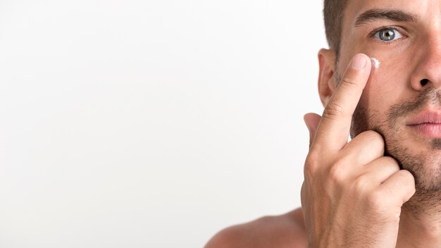 Half face of shirtless young man applying cream on his face against white background