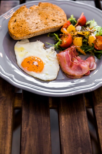 Half fried egg; toast; salad; bacon on gray ceramic plate on wooden table