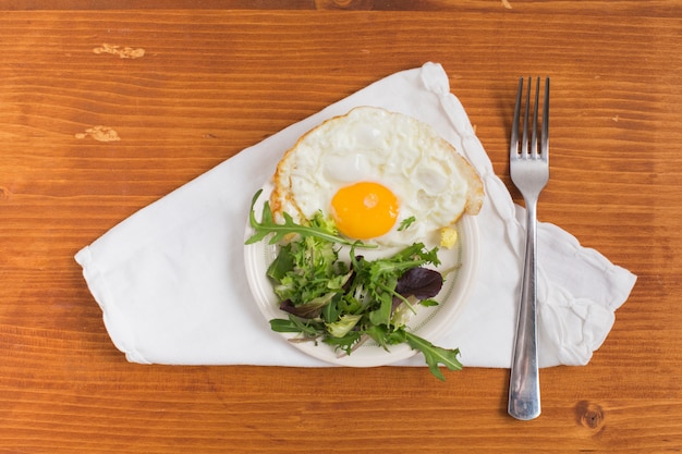 Half fried egg with salad on plate and fork over the white napkin against wooden textured backdrop