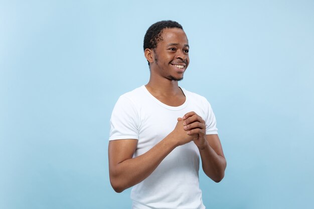 Half-length close up portrait of young african-american male model in white shirt on blue background. Human emotions, facial expression, ad concept. Doubts, asking, showing uncertainty, smiling.