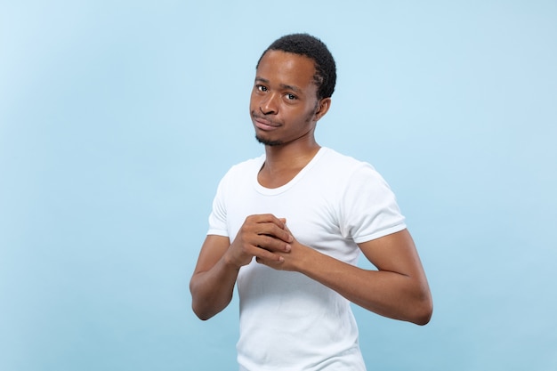 Half-length close up portrait of young african-american male model in white shirt on blue background. Human emotions, facial expression, ad concept. Doubts, asking, showing uncertainty, thoughtful.