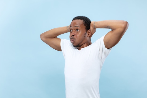 Half-length close up portrait of young african-american male model in white shirt on blue space. Human emotions, facial expression, ad or betting concept