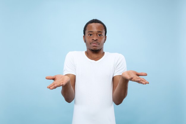 青いスペースに白いシャツを着た若いアフリカ系アメリカ人男性モデルのハーフレングスのクローズアップの肖像画。人間の感情、表情、広告コンセプト