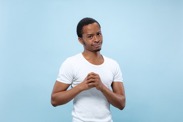 Half-length close up portrait of young african-american male model in white shirt on blue wall. Human emotions, facial expression, ad concept. Doubts, asking, showing uncertainty, thoughtful.
