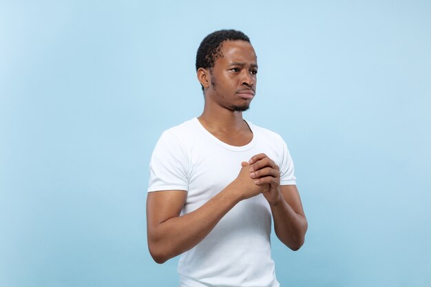 青い壁に白いシャツを着た若いアフリカ系アメリカ人男性モデルのハーフレングスのクローズアップの肖像画。人間の感情、顔の表情、広告のコンセプト。疑い、尋ね、不確実性を示し、思慮深い。