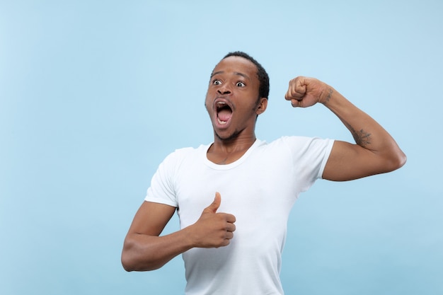 Half-length close up portrait of young african-american man in white shirt on blue background. Human emotions, facial expression, ad, concept. Celebrating, wondered, astonished, shocked, crazy happy.