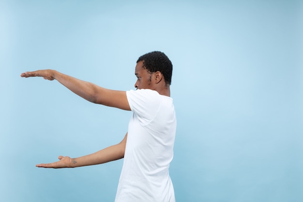 Half-length close up portrait of young african-american man in white shirt on blue background. Human emotions, facial expression, ad concept. Holding an empty bar, copyspace for your text.