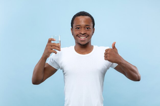Half-length close up portrait of young african-american man in white shirt on blue background. Human emotions, facial expression, ad concept. Holding a glass and drinking water.