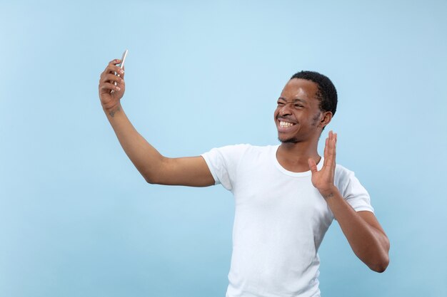 Half-length close up portrait of young african-american man in white shirt on blue background. Human emotions, facial expression, ad concept. Making selfie or content for social media, vlog.