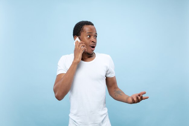 Half-length close up portrait of young african-american man in white shirt on blue background. Human emotions, facial expression, ad concept. Talking on the phone, holding a smartphone.