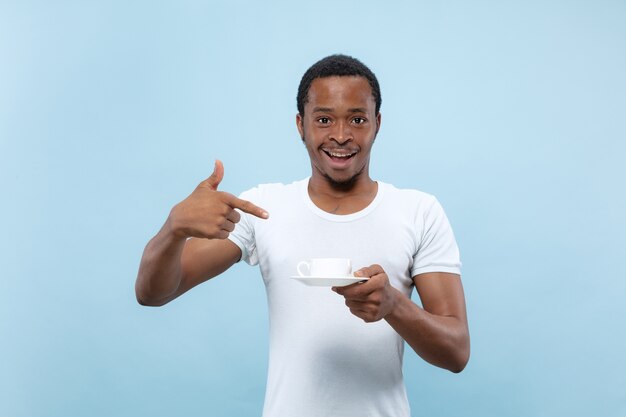 Half-length close up portrait of young african-american man in white shirt on blue background. Human emotions, facial expression, ad, sales, concept. Enjoying, drinking coffee, smiling. Pointing on.