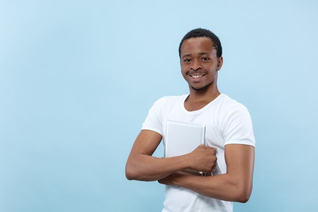 青の背景に白いシャツを着た若いアフリカ系アメリカ人の男性の半分の長さのクローズアップの肖像画。人間の感情、表情、広告、販売コンセプト。タブレットを持って笑顔。幸せそうだ。