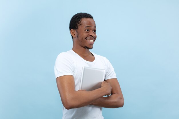 青いスペースに白いシャツを着た若いアフリカ系アメリカ人男性のハーフレングスのクローズアップの肖像画。タブレットを持って笑顔
