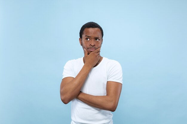 Half-length close up portrait of young african-american man in white shirt on blue space. Human emotions, facial expression, ad concept