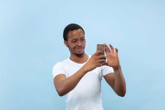 Half-length close up portrait of young african-american man in white shirt on blue space. Human emotions, facial expression, ad concept