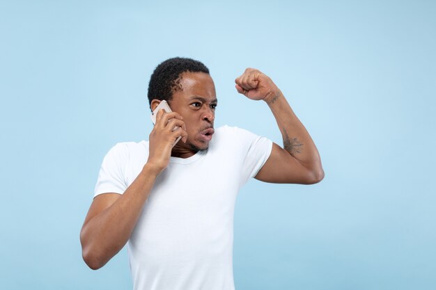 Half-length close up portrait of young african-american man in white shirt on blue space. Human emotions, facial expression, ad concept