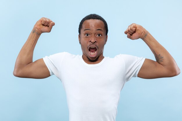 Half-length close up portrait of young african-american man in white shirt on blue space. Human emotions, facial expression, ad, concept