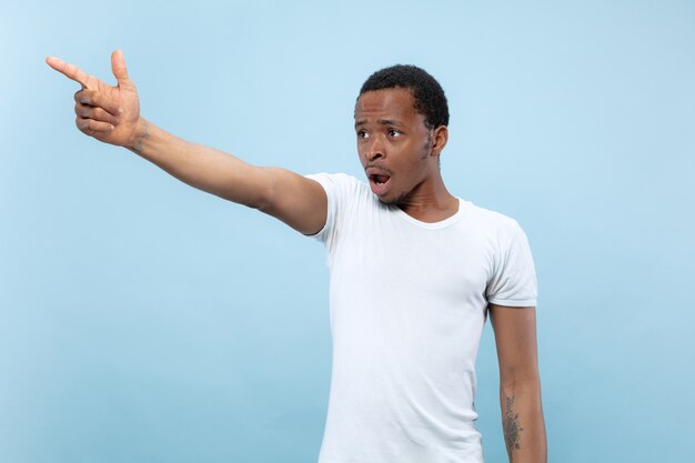 Half-length close up portrait of young african-american man in white shirt on blue space. Human emotions, facial expression, ad, sales concept. Pointing, choosing, astonished