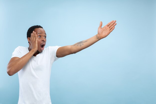 Half-length close up portrait of young african-american man in white shirt on blue space. Human emotions, facial expression, ad, sales concept. Pointing, choosing, astonished