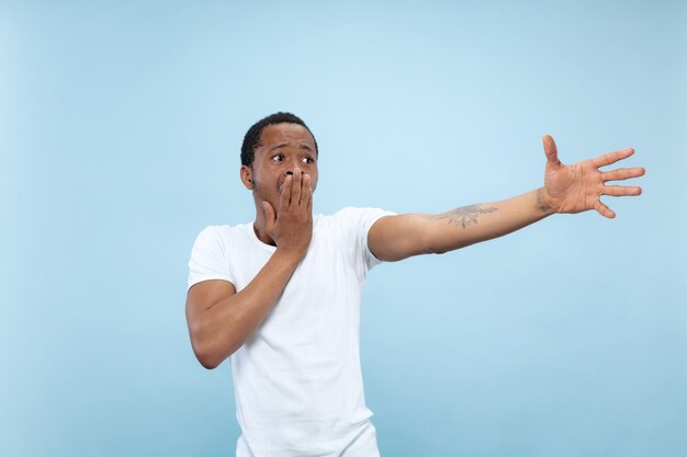 Half-length close up portrait of young african-american man in white shirt on blue space. Human emotions, facial expression, ad, sales concept. Pointing, choosing, astonished