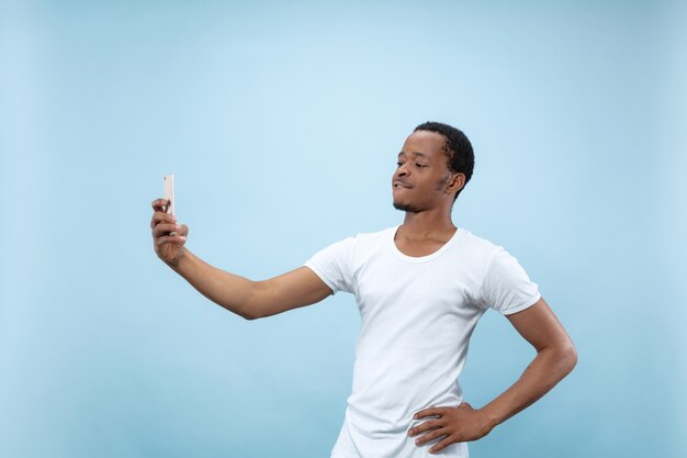 Half-length close up portrait of young african-american man in white shirt on blue wall. Human emotions, facial expression, ad concept. Making selfie or content for social media, vlog.