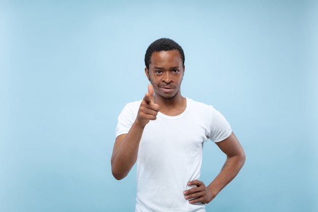 Поясной конец вверх по портрету молодого афро-американского человека в белой рубашке на голубой стене. Человеческие эмоции, выражение лица, концепция рекламы. Указывая, выбирая и улыбаясь. Copyspace.