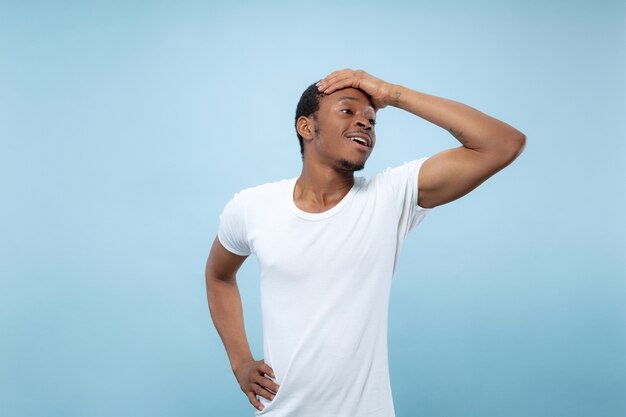 Half-length close up portrait of young african-american man in white shirt on blue wall. Human emotions, facial expression, ad concept. Sales, betting. Looks excited, astonished, crazy happy.