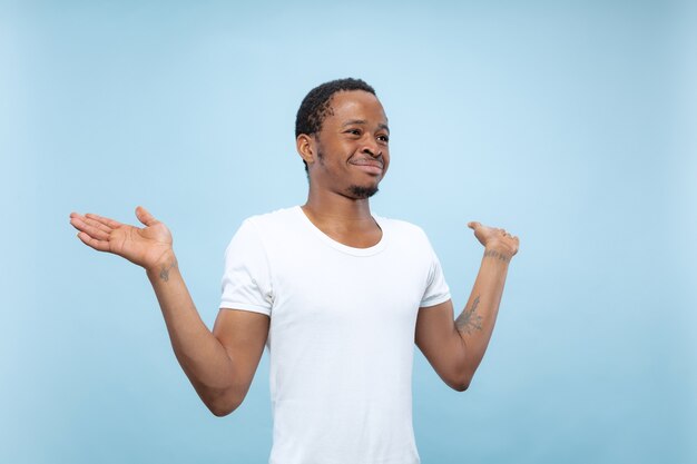 Half-length close up portrait of young african-american man in white shirt on blue wall. Human emotions, facial expression, ad concept. Showing empty bar, pointing, choosing, inviting.