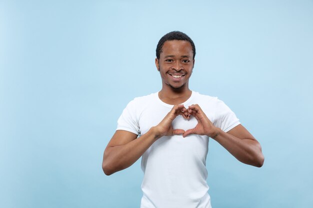 Half-length close up portrait of young african-american man in white shirt on blue wall. Human emotions, facial expression, ad concept. Showing the sign of a heart by his hands, smiling.
