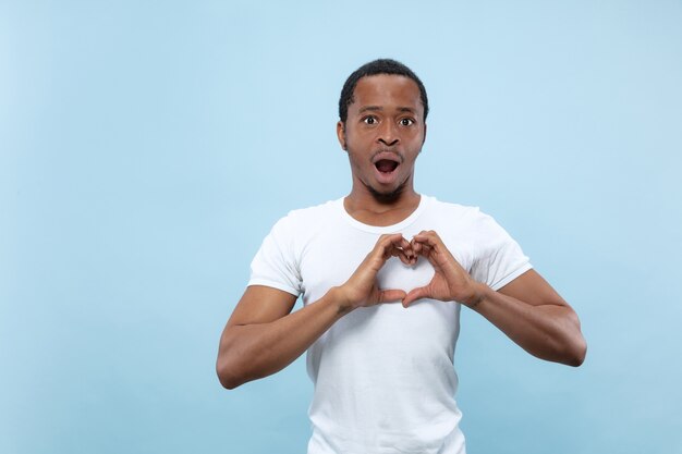 Half-length close up portrait of young african-american man in white shirt on blue wall. Human emotions, facial expression, ad concept. Showing the sign oh a heart, astonished.