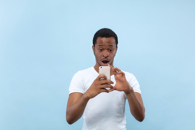 Half-length close up portrait of young african-american man in white shirt on blue wall. Human emotions, facial expression, ad concept. Taking a photo or vlog content on his smartphone.