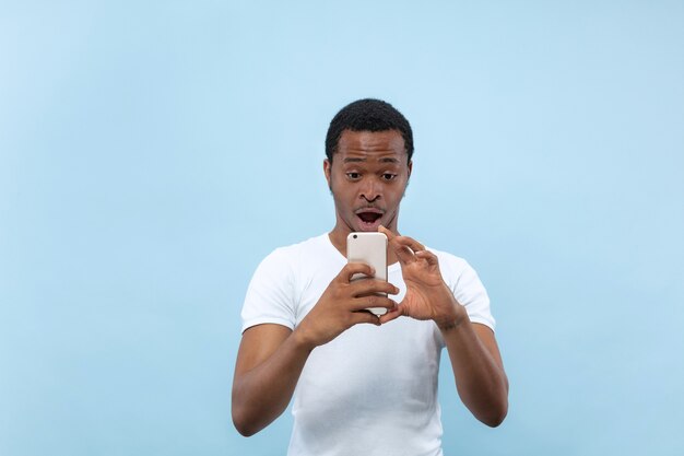 青い壁に白いシャツを着た若いアフリカ系アメリカ人男性のハーフレングスのクローズアップの肖像画。人間の感情、顔の表情、広告のコンセプト。彼のスマートフォンで写真やvlogのコンテンツを撮る。