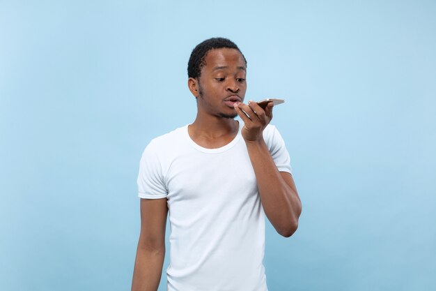 Half-length close up portrait of young african-american man in white shirt on blue wall. Human emotions, facial expression, ad concept. Talking on the smartphone or recording a voice message.