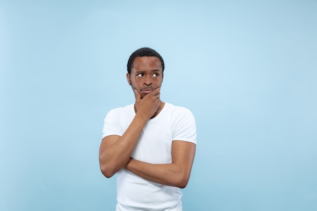 Half-length close up portrait of young african-american man in white shirt on blue wall. Human emotions, facial expression, ad concept. Thoughtful, thinking covering face with his hands.