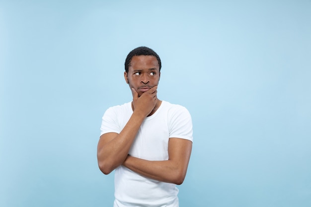 青い壁に白いシャツを着た若いアフリカ系アメリカ人男性のハーフレングスのクローズアップの肖像画。人間の感情、顔の表情、広告のコンセプト。思いやり、手で顔を覆うことを考えています。