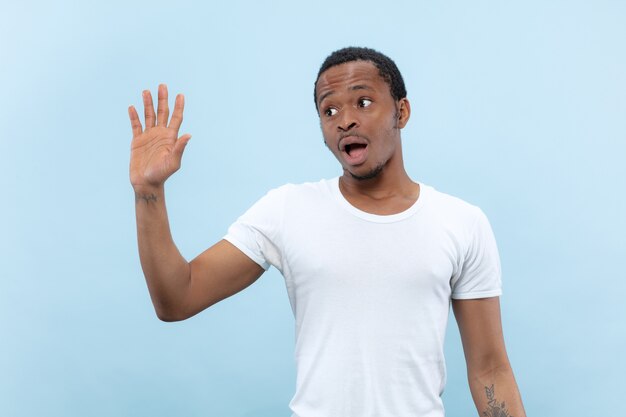 Half-length close up portrait of young african-american man in white shirt on blue wall. Human emotions, facial expression, ad, sales concept. Meeting somebody, greeting, inviting.