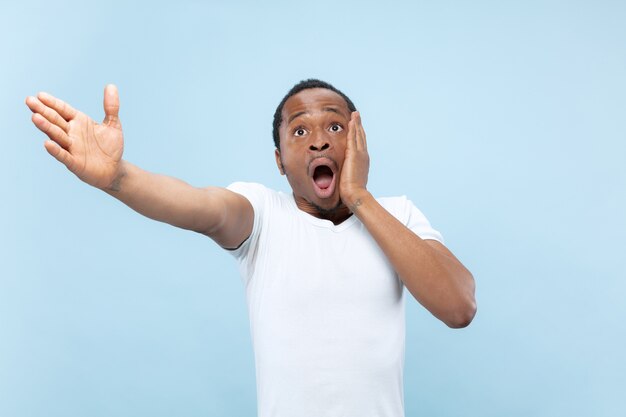 Half-length close up portrait of young african-american man in white shirt on blue wall. Human emotions, facial expression, ad, sales concept. Pointing, choosing, astonished. Copyspace.