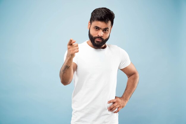 Half-length close up portrait of young hindoo man in white shirt on blue background. Human emotions, facial expression, ad concept. Negative space. Showing empty bar, pointing, choosing, inviting.