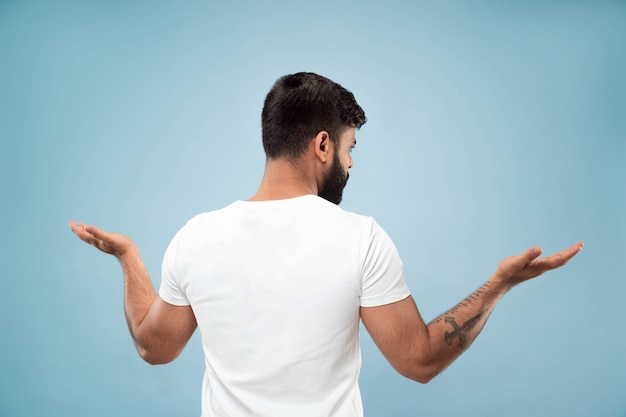 Half-length close up portrait of young hindoo man in white shirt on blue background. Human emotions, facial expression, ad concept. Negative space. Showing empty bar, pointing, choosing, inviting.