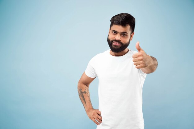 Half-length close up portrait of young hindoo man in white shirt on blue background. Human emotions, facial expression, ad concept. Negative space. Showing the sign of OK, nice, great. Smiling.