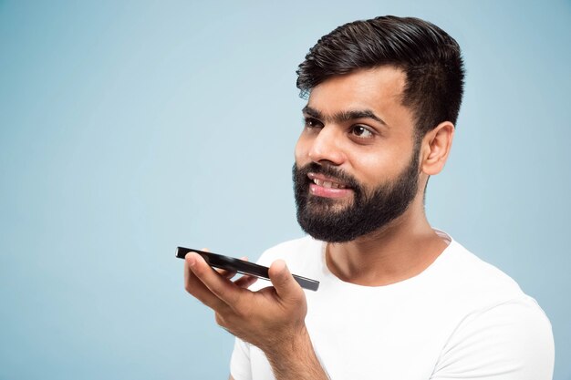 青の背景に白いシャツを着た若いヒンドゥー教の男の肖像画の半分のクローズアップ。人間の感情、顔の表情、広告のコンセプト。ネガティブスペース。携帯電話で話し、音声メッセージを録音します。
