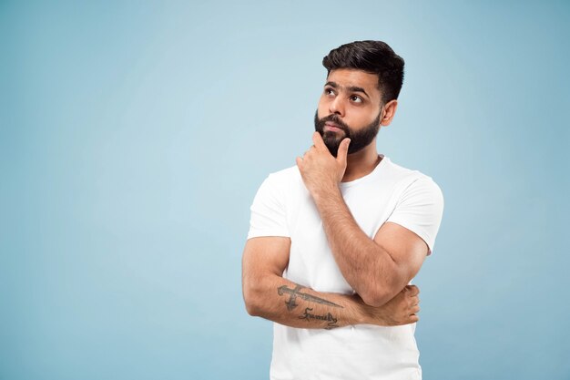 Поясной конец вверх по портрету молодого индусского человека в белой рубашке на синем фоне. Человеческие эмоции, выражение лица, концепция рекламы. Негативное пространство. Думал, держась за бороду. Выбор.