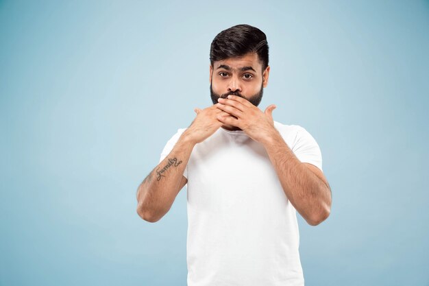 Half-length close up portrait of young hindoo man in white shirt on blue background. Human emotions, facial expression, sales, ad concept. Negative space. Covering his face with his hands.